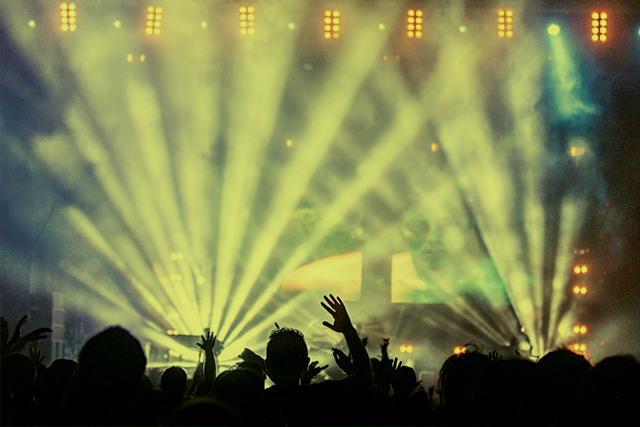 Graphite pencil sketch of a festival crowd with raised hands in front of stage lights, soft tonal shading